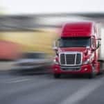 A red semi truck in focus driving on a road with everything around the truck appearing blurry or out of focus.