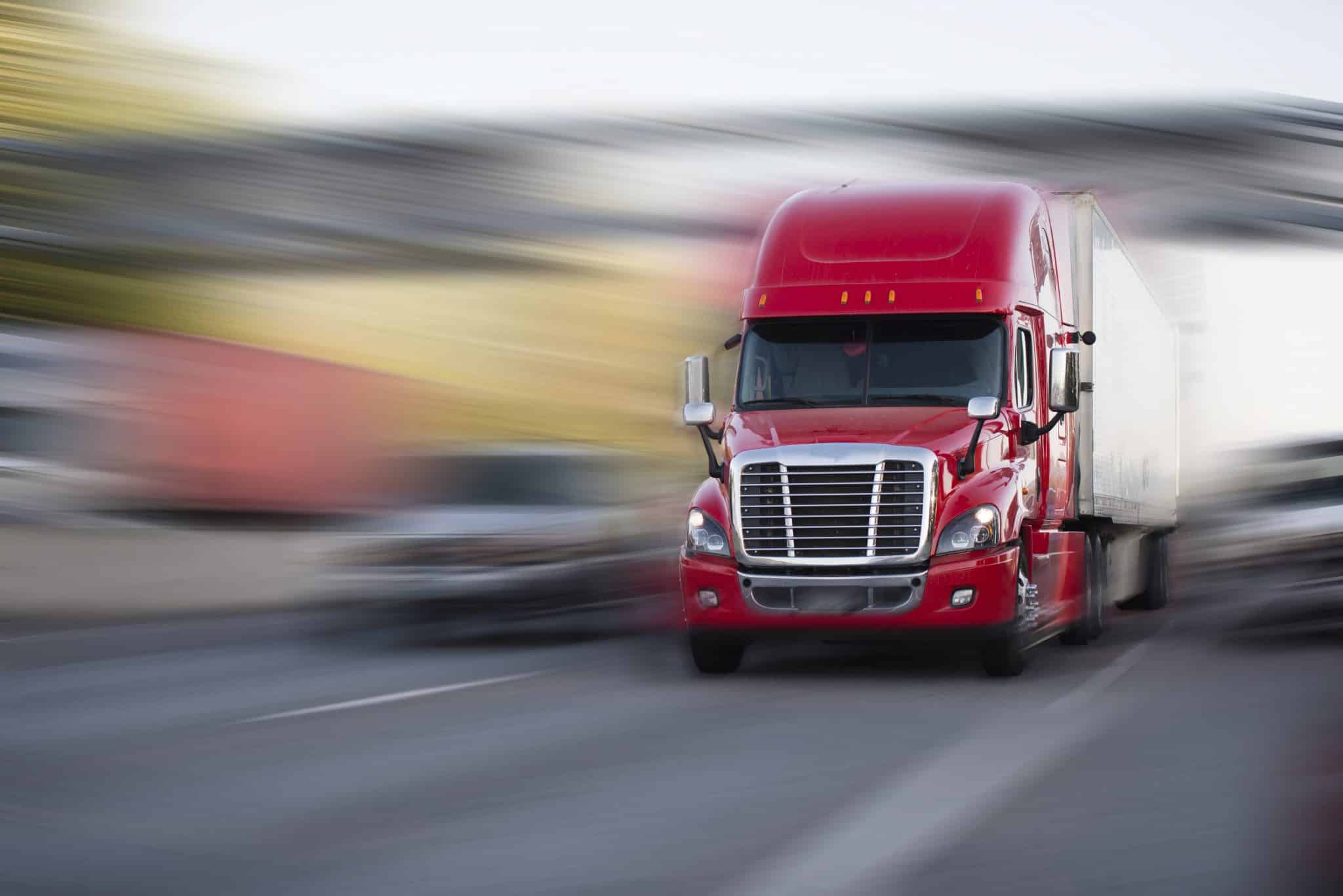 A red semi truck in focus driving on a road with everything around the truck appearing blurry or out of focus.