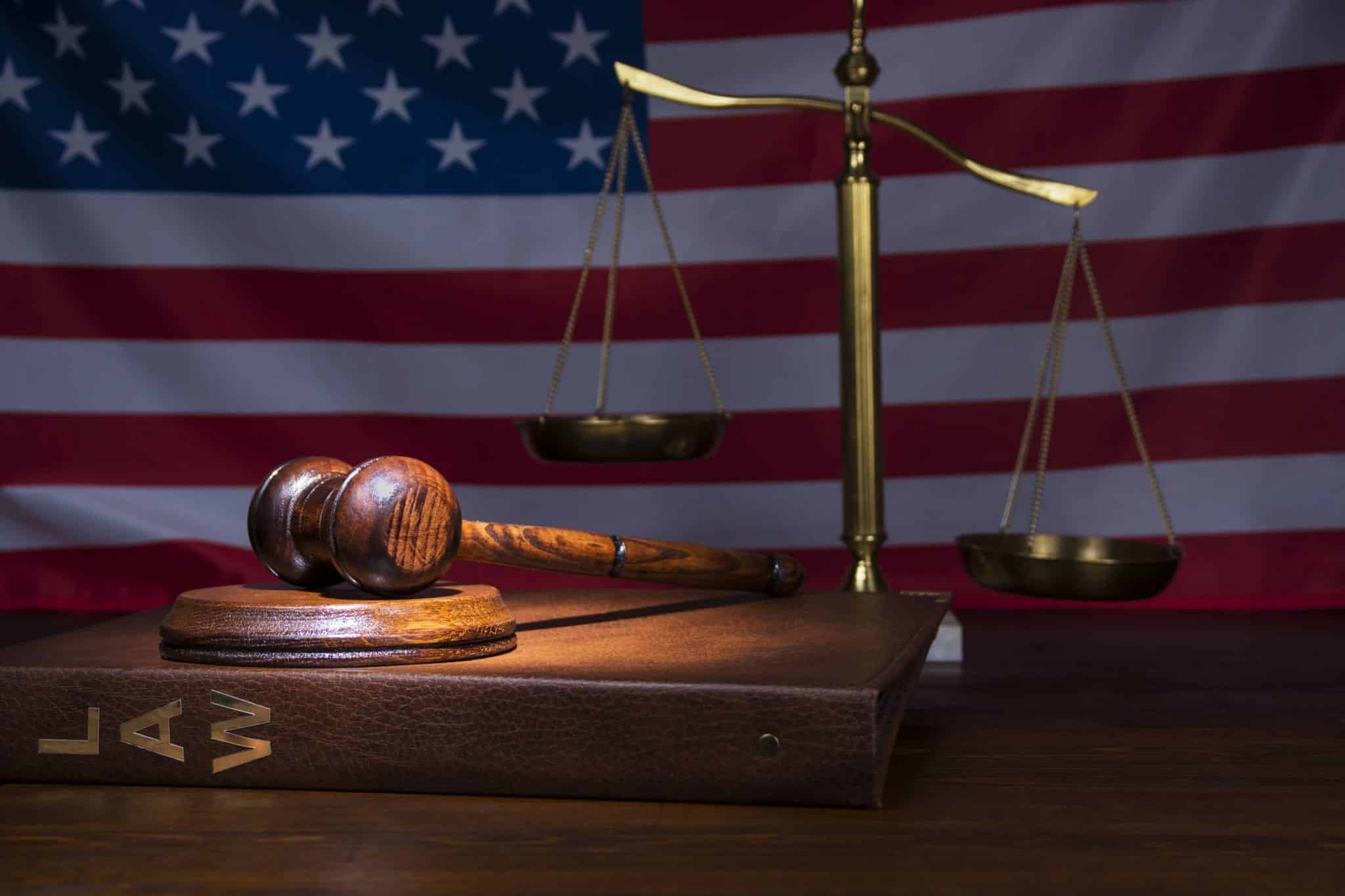 A brown binder with gold letters spelling LAW laying on a wood table with a wooden gavel resting on top of it in front of a shiny gold scale with an American flag in the background.