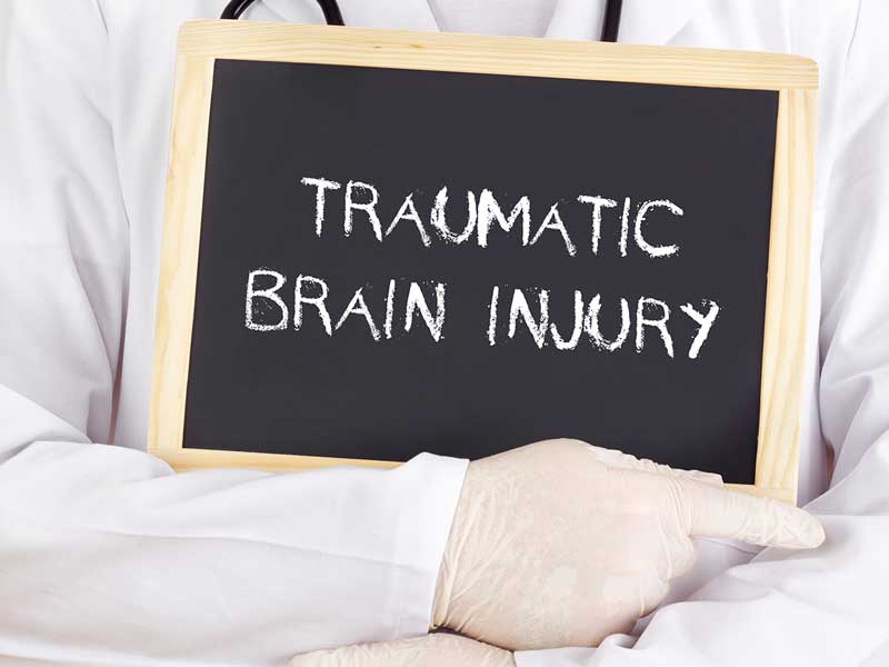 A close-up of someone's chest wearing a white coat and white surgical gloves with a stethoscope around their neck, holding a black chalkboard with the words TRAUMATIC BRAIN INJURY written on it.