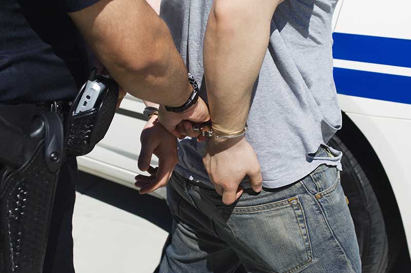 Close-up of back of man in handcuffs being led by a police officer to a cop car.