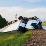 A wrecked, overturned white tractor trailer truck in the green grass of a ditch on the side of a country road.