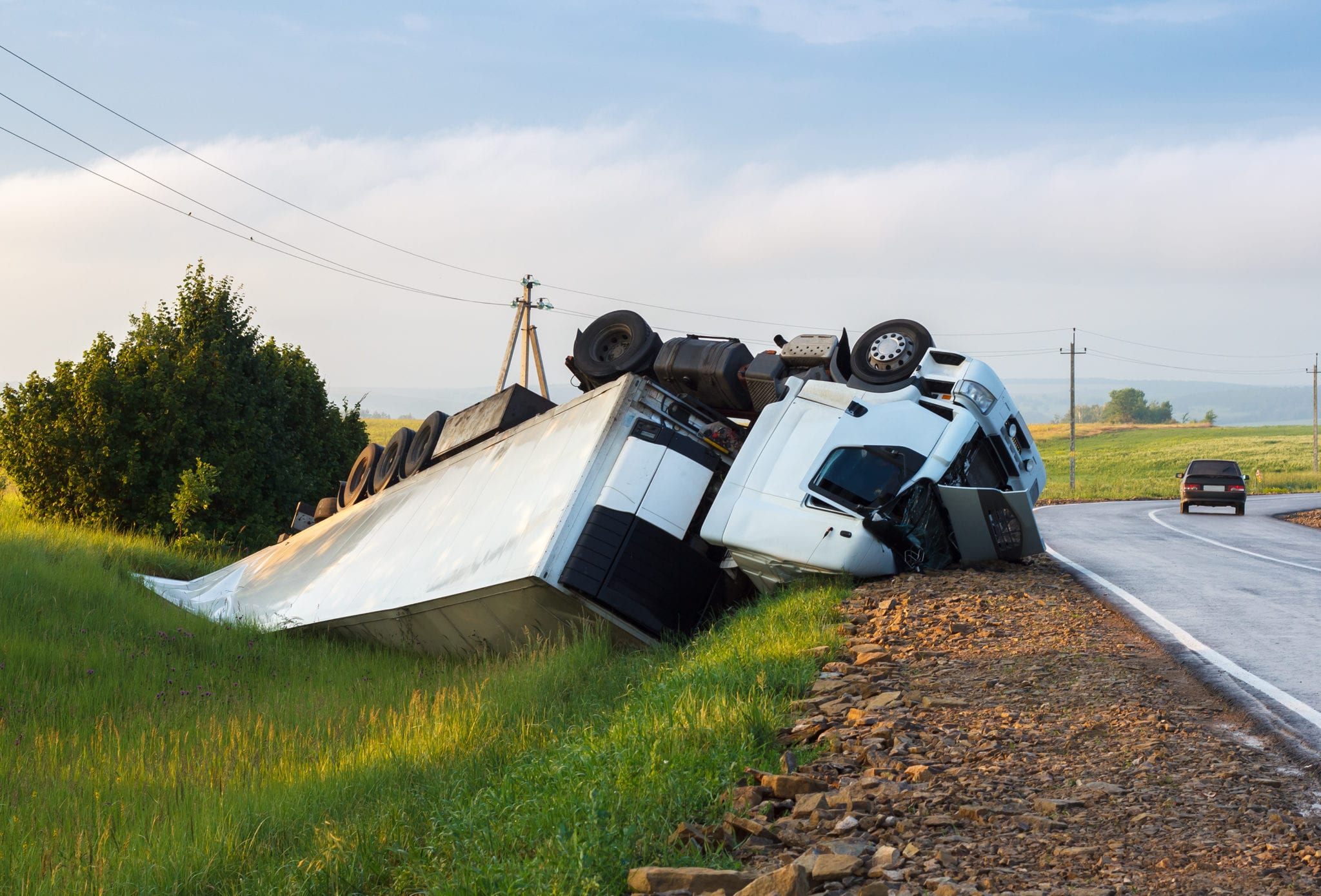 A wrecked, overturned white tractor trailer truck in the green grass of a ditch on the side of a country road.