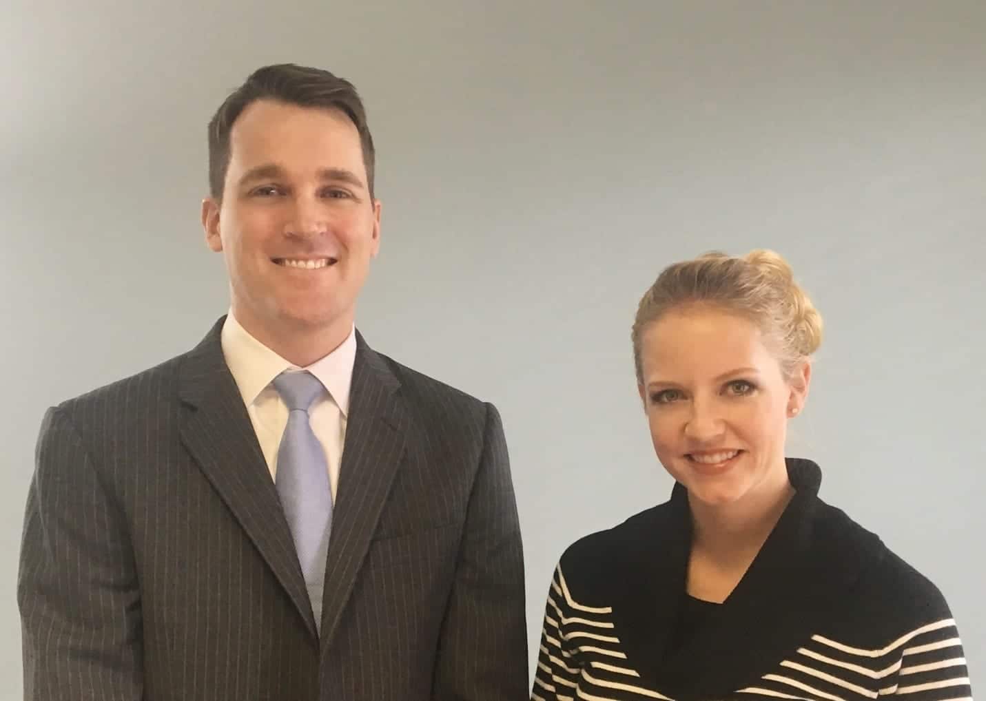 A tall man in a gray business suit with a blue tie standing besides a blonde woman wearing a black and white striped shirt.