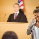 A blurry background of a seated judge in front of an american flag in a courtroom with a woman in the foreground in clear focus looking down, pressing her fingers against her forehead, as she walks away from a witness stand.