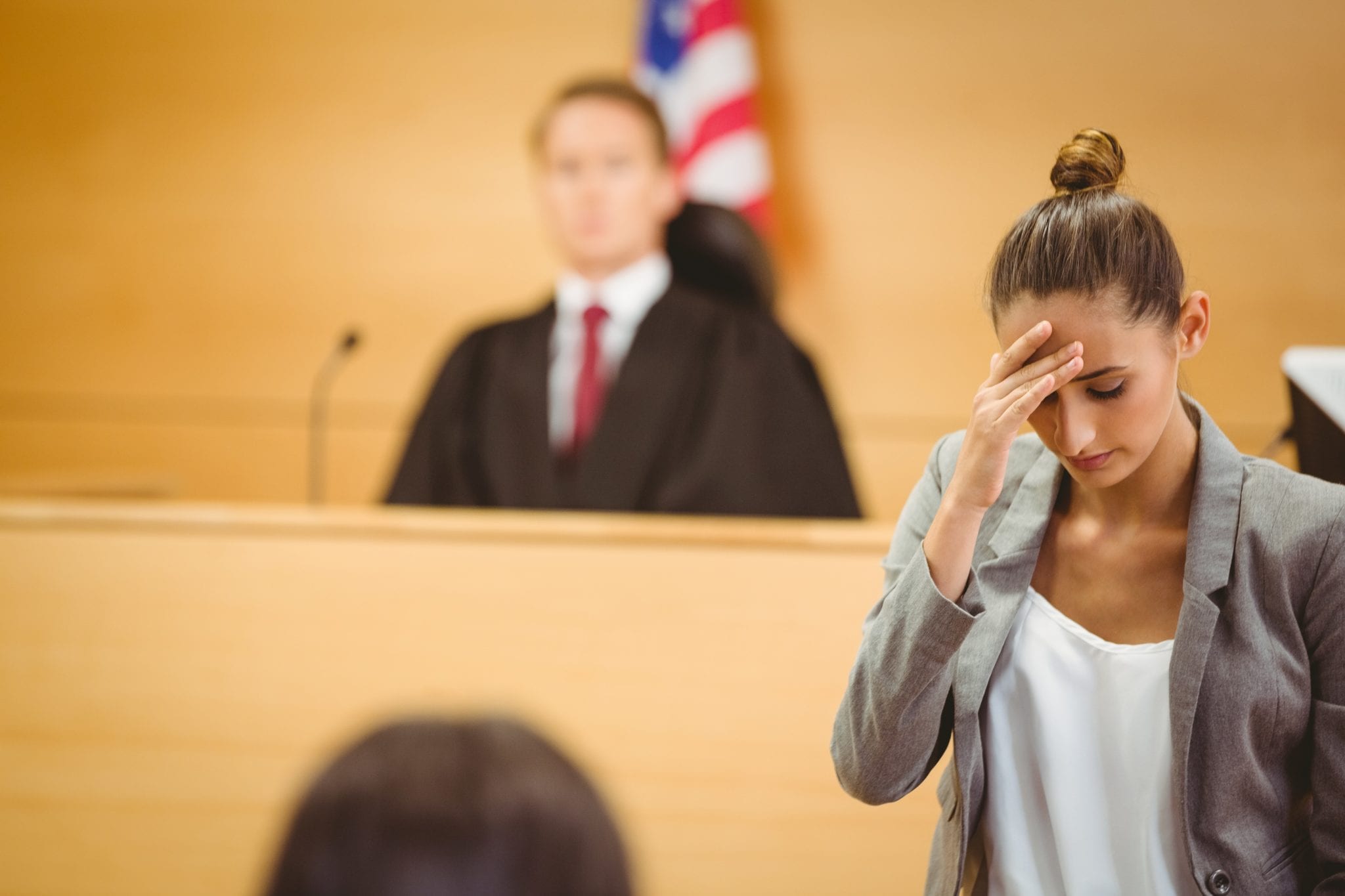 A blurry background of a seated judge in front of an american flag in a courtroom with a woman in the foreground in clear focus looking down, pressing her fingers against her forehead, as she walks away from a witness stand.