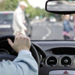 The interior of a vehicle showing the steering wheel and dashboard with driver's hand on the wheel and a blurry view through the windshield of an adult and two kids walking across the road.