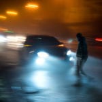 A blurry image at night of traffic on a busy road and a car in focus about to hit a man crossing the street.