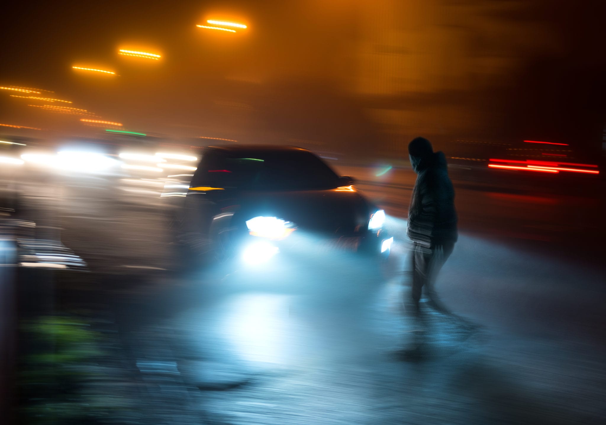 A blurry image at night of traffic on a busy road and a car in focus about to hit a man crossing the street.