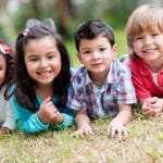 A close-up of four youg children, two boys and two girls laying in the grass on there stomachs, proped up on their elbows.