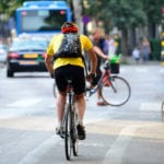 A cyclist wearing a yellow t-shirt, red helmet and black bikershorts and backpack riding in a bike lane on along a city street with a bus and cars in the blurred background.