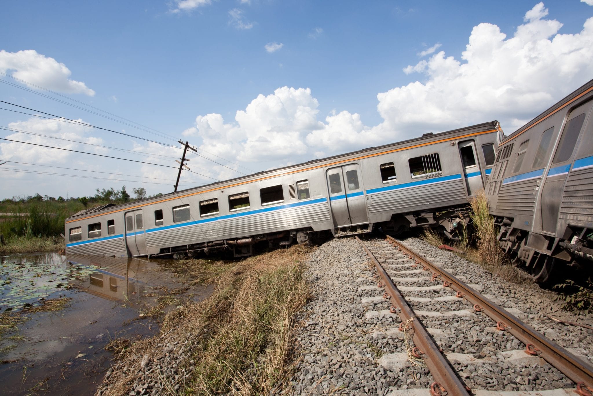 A badly derailed silver passenger train with the back portion partially submerged in a large ditch full of muddy water under a blue sky with white fluffy clouds.