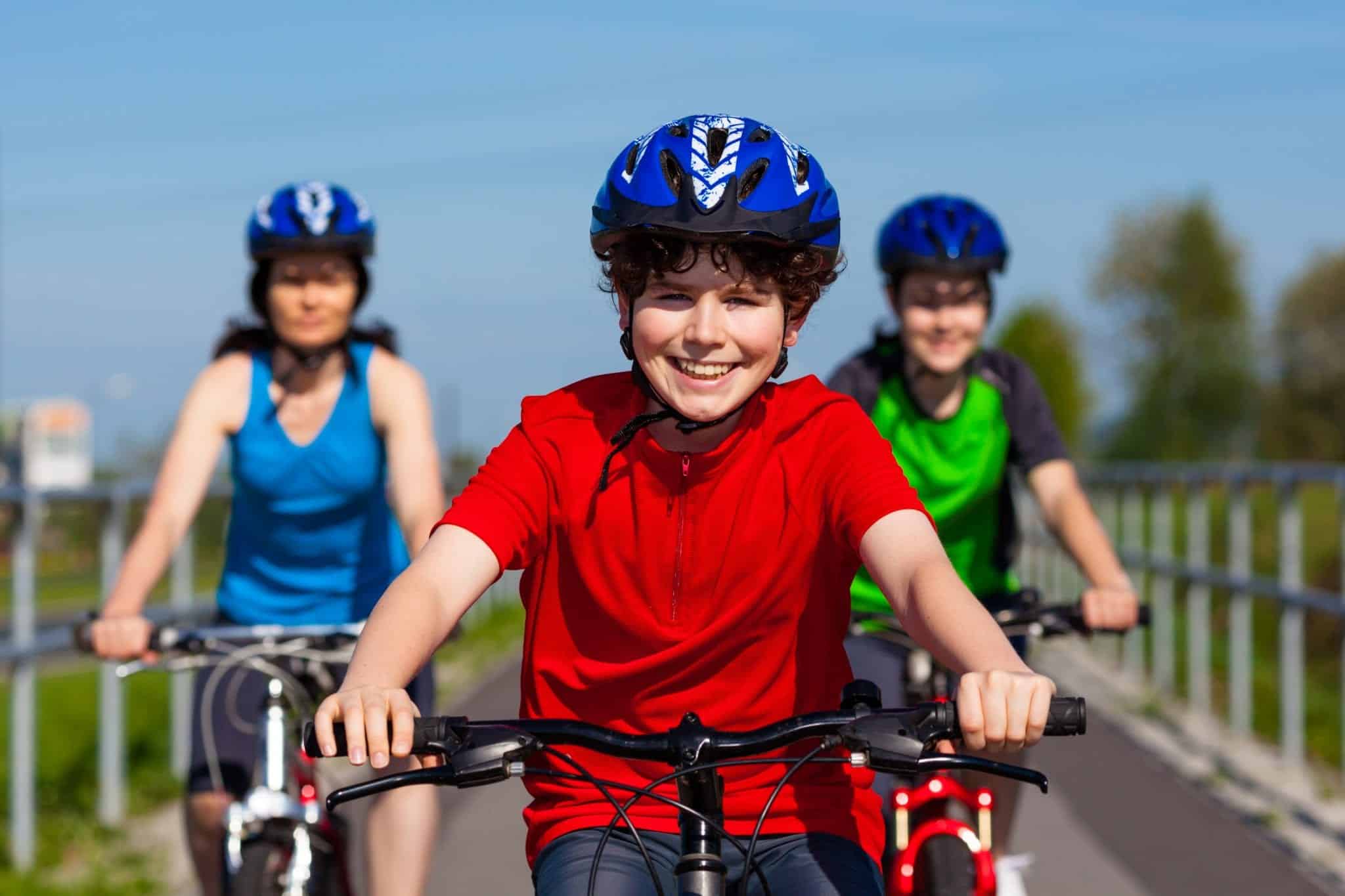 A boy wearing a bright red shirt and blue helmet riding a bike with a woman and boy blurred in the background, wearing blue and neon green shirts respectively, also riding bikes on a bike path.