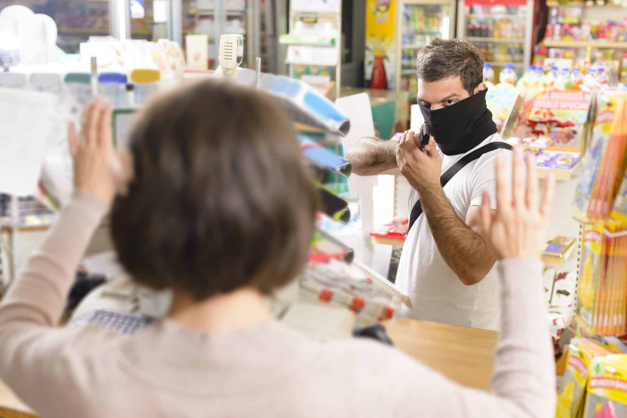 The back of a woman with short brown hair, standing inside a store, holding both hands up, with a man facing her, wearing a black mask, pointing a gun at her.