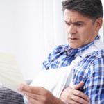 A middle aged man wearing a blue and white plaid shirt with his right arm in a white sling holding up a piece of paper with a look of concern on his face.