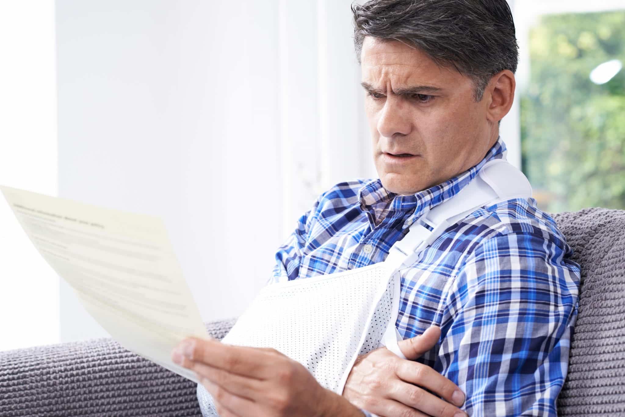 A middle aged man wearing a blue and white plaid shirt with his right arm in a white sling holding up a piece of paper with a look of concern on his face.