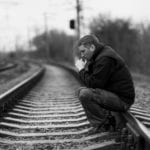 A black and white photo of a worried man sitting on railroad tracks with his hands folded near his chin.