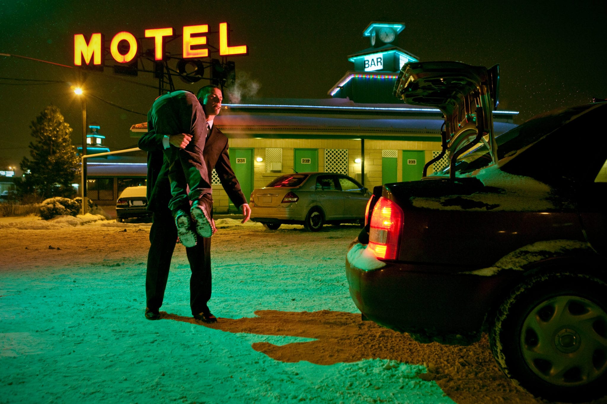 A man in a black suit carrying a passed out man over his shoulder at night, away from a motel, towards an open trunk of a car.