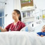 A young child wearing a blue hospital gown, sleeping in a hospital bed with a nurse in red scrubs and a woman wearing a teal sweater, standing in the background, near the hospital bed having a conversation.