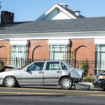 The side view of a brick building and three cars in a line, along the road, that have been involved in a car wreck with the first car having it's rearend smashed, the middle car having it's front and rear end smashed and the back car has it's front end smashed.