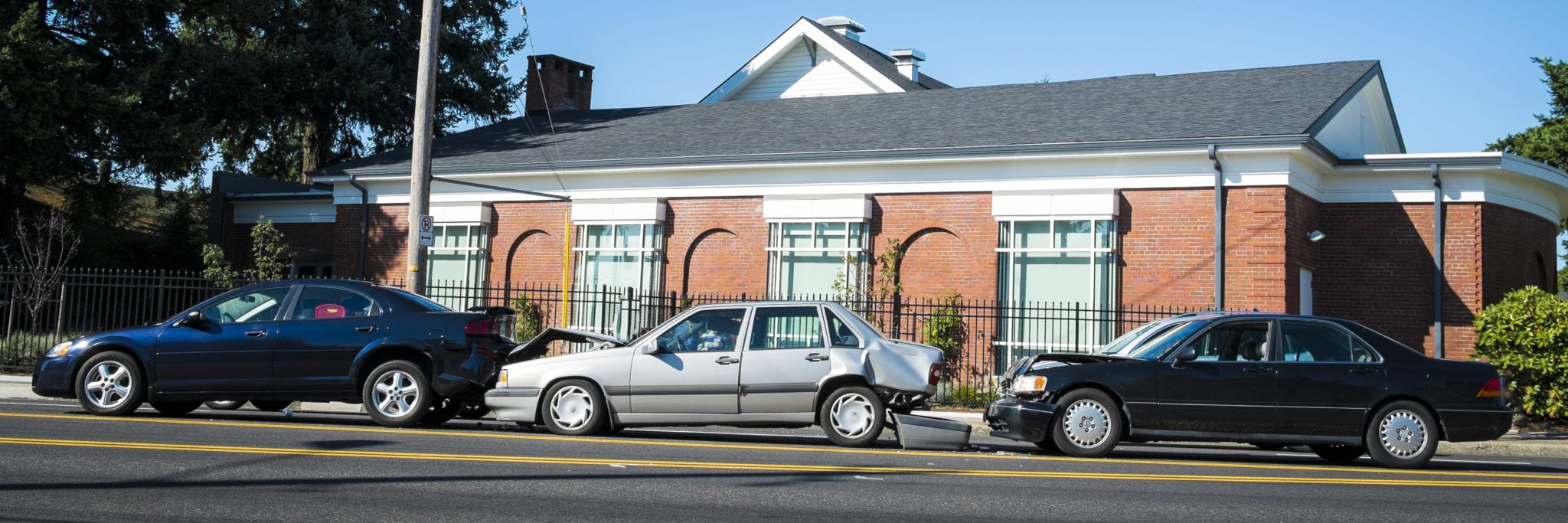 The side view of a brick building and three cars in a line, along the road, that have been involved in a car wreck with the first car having it's rearend smashed, the middle car having it's front and rear end smashed and the back car has it's front end smashed.