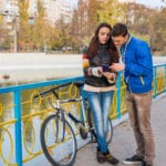 A man and woman looking at a phone screen together while standing near a bicycle, propped against a railing with a body of water in the background.