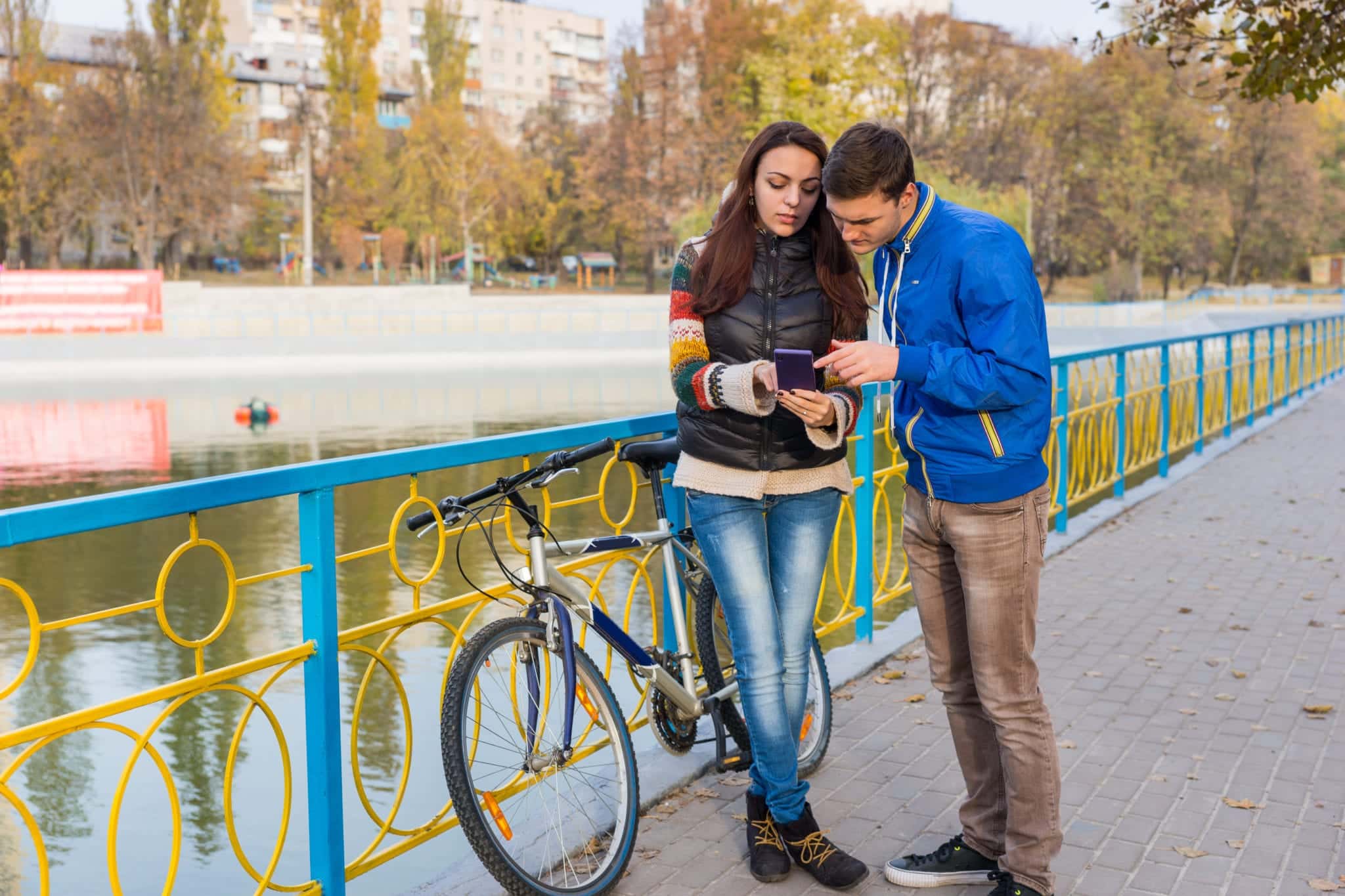 A man and woman looking at a phone screen together while standing near a bicycle, propped against a railing with a body of water in the background.