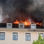 A tan colored house with orange fire coming out of the roof and a dark cloud of smoke.