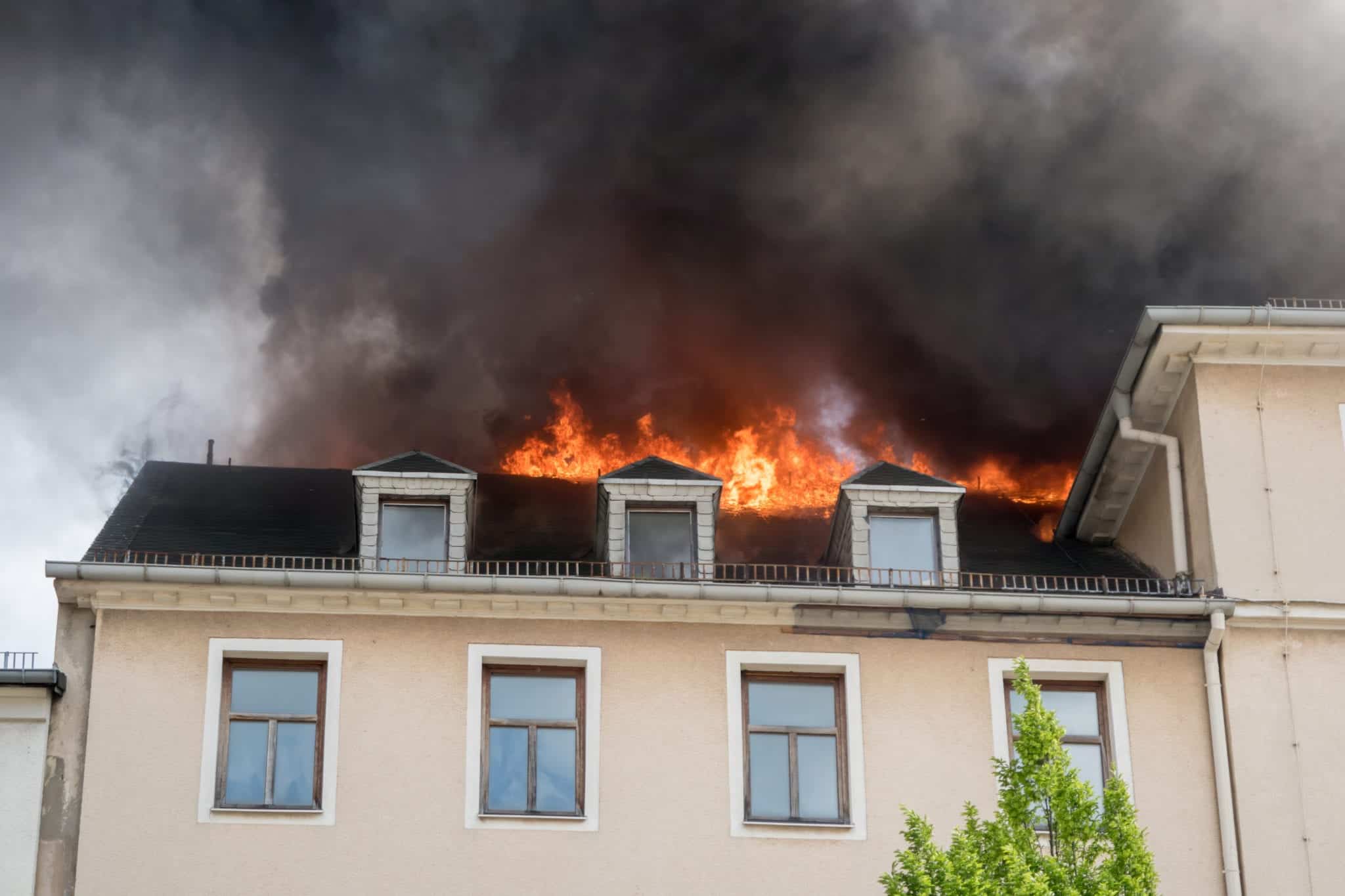 A tan colored house with orange fire coming out of the roof and a dark cloud of smoke.