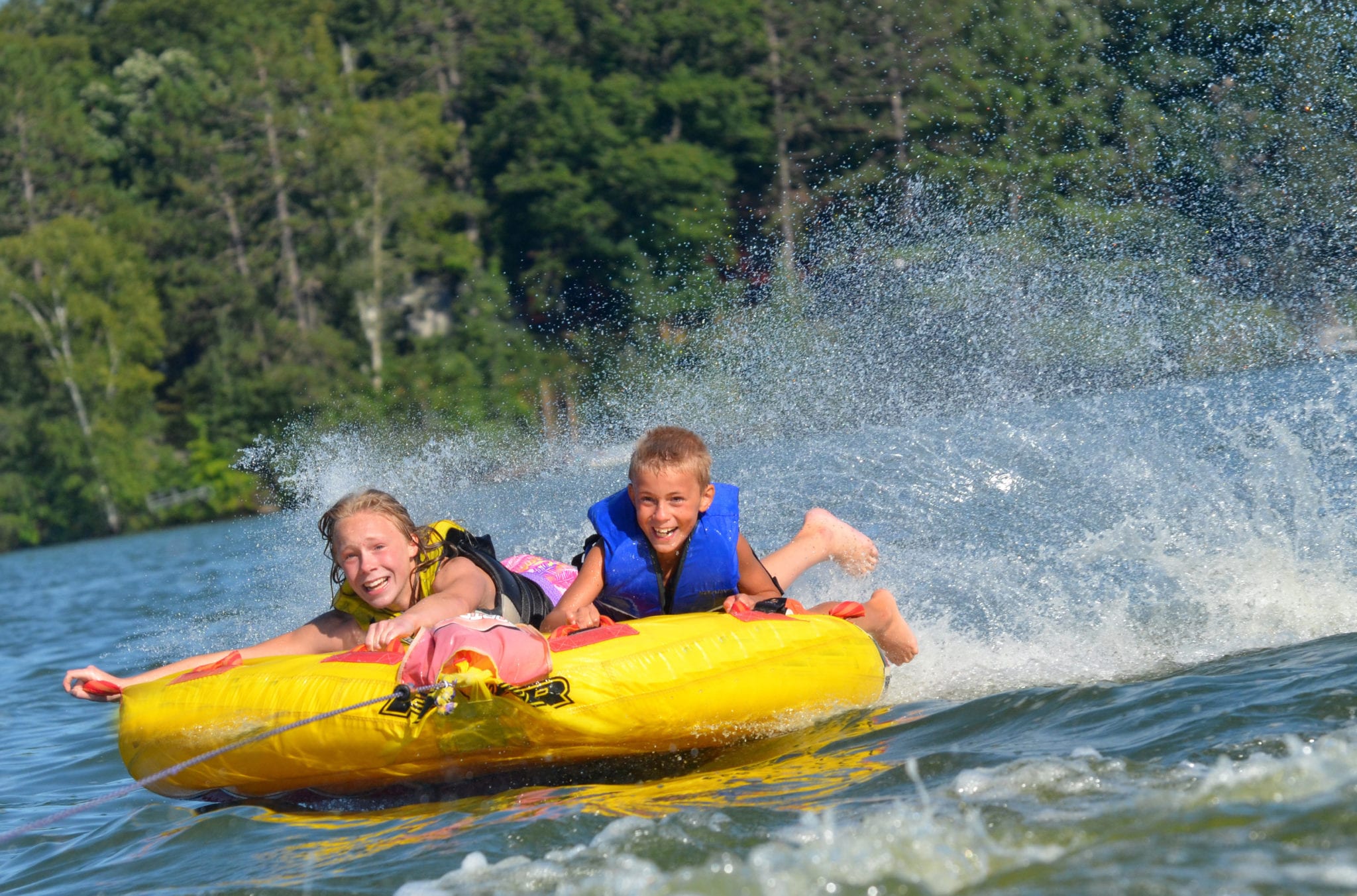 Two kids on a yellow tube being pulled by a boat on a lake.