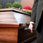 A close-up of a man, from his waist to just above his knees, wearing a dark gray suit holding the silver handle of a brown wooden casket.