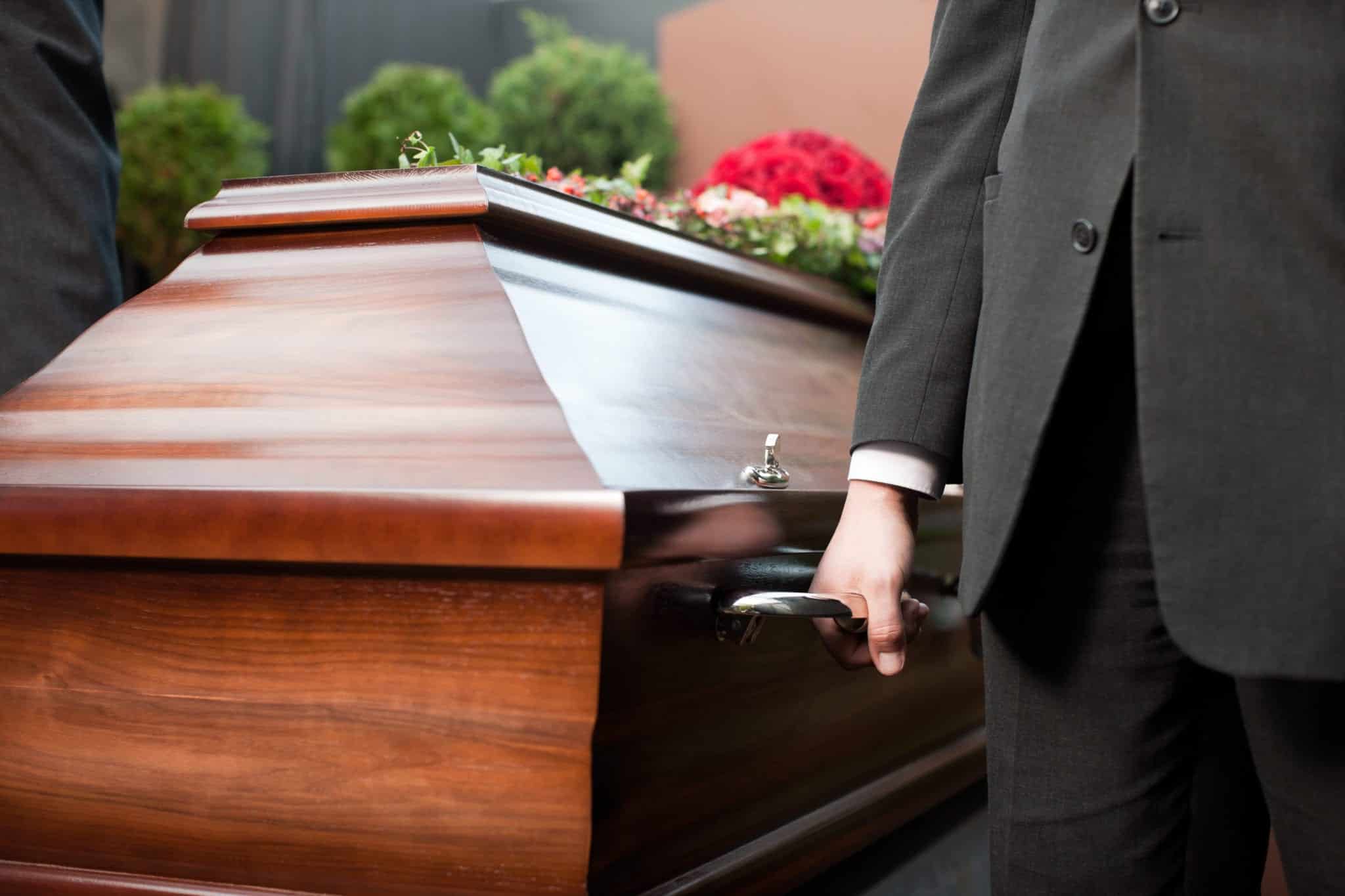 A close-up of a man, from his waist to just above his knees, wearing a dark gray suit holding the silver handle of a brown wooden casket.