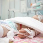 An injured child laying on a hospital bed, an IV in their left hand, tubes coming from their mouth, white cloths securing the child to the bed and a blurry background of hospital monitors.