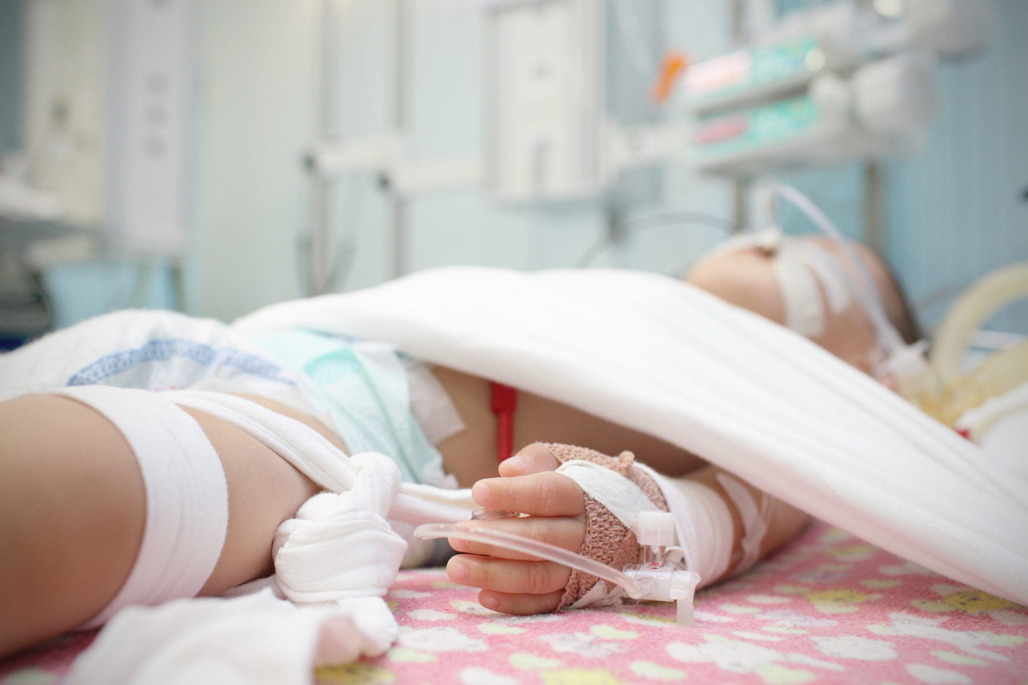 An injured child laying on a hospital bed, an IV in their left hand, tubes coming from their mouth, white cloths securing the child to the bed and a blurry background of hospital monitors.