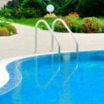 The blue water of an outdoor, inground pool with metal railings for stairs, surrounded by a tan concrete pool deck and green landscaping in the background.