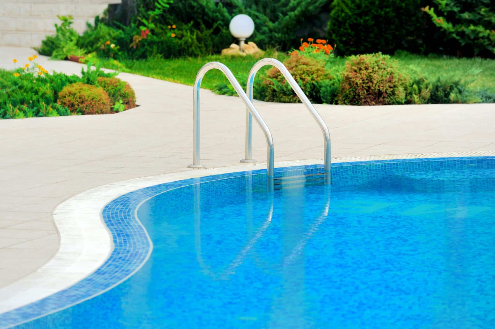 The blue water of an outdoor, inground pool with metal railings for stairs, surrounded by a tan concrete pool deck and green landscaping in the background.