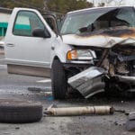 An extremely damaged front end view of a white truck with a burnt, smashed in hood, with it's front left tire laying on the ground in front of it.