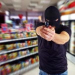 A man wearing a black t-shirt and ski mask, standing in a convenience store, using both hands to point a gun at the camera.