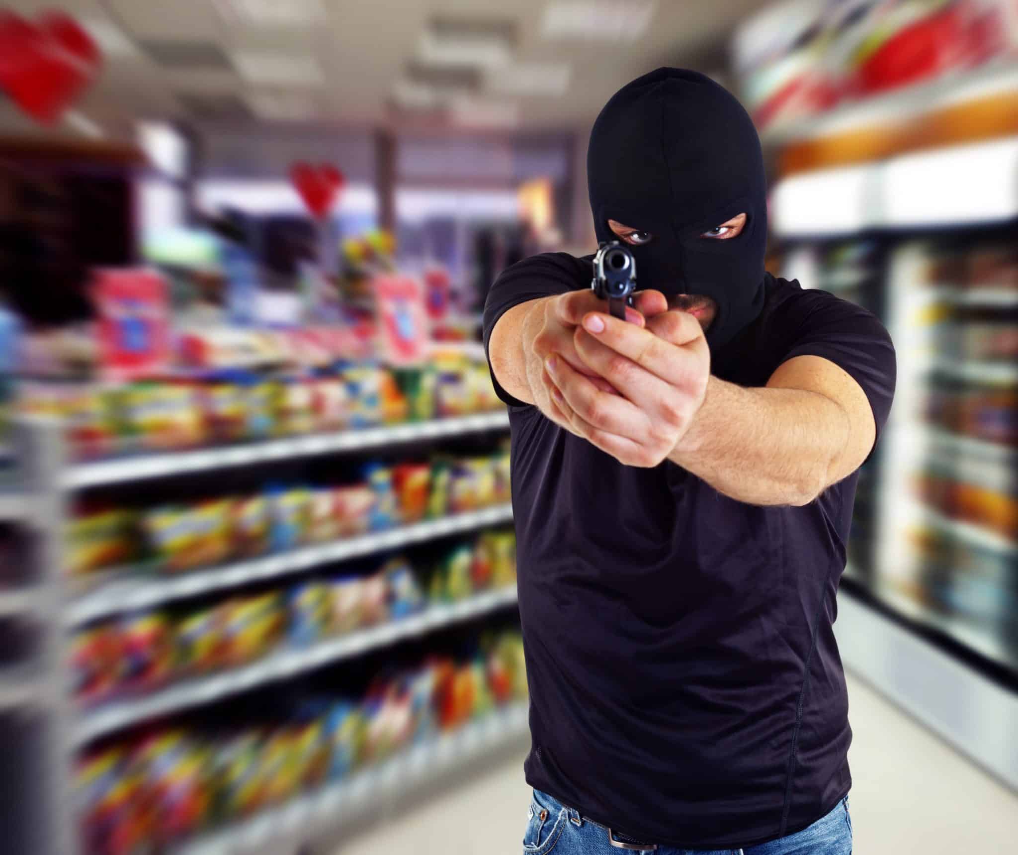 A man wearing a black t-shirt and ski mask, standing in a convenience store, using both hands to point a gun at the camera.