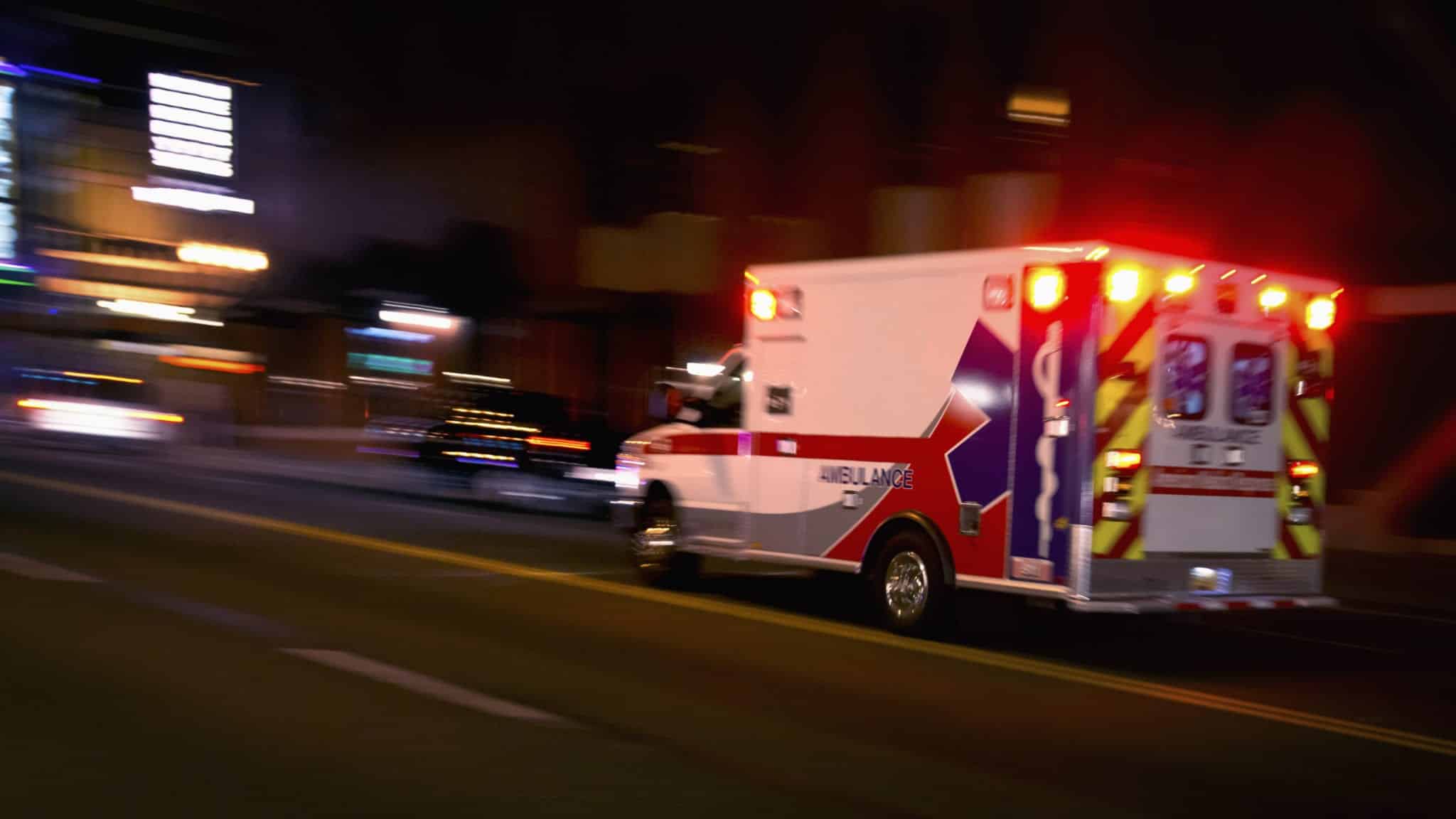 A blurry view of an ambulance with it's lights on driving up the road at night.