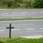 A divided multi-lane highway with green grass on each side, as well as the center median, and a wooden memoral cross standing in the grass near the road.