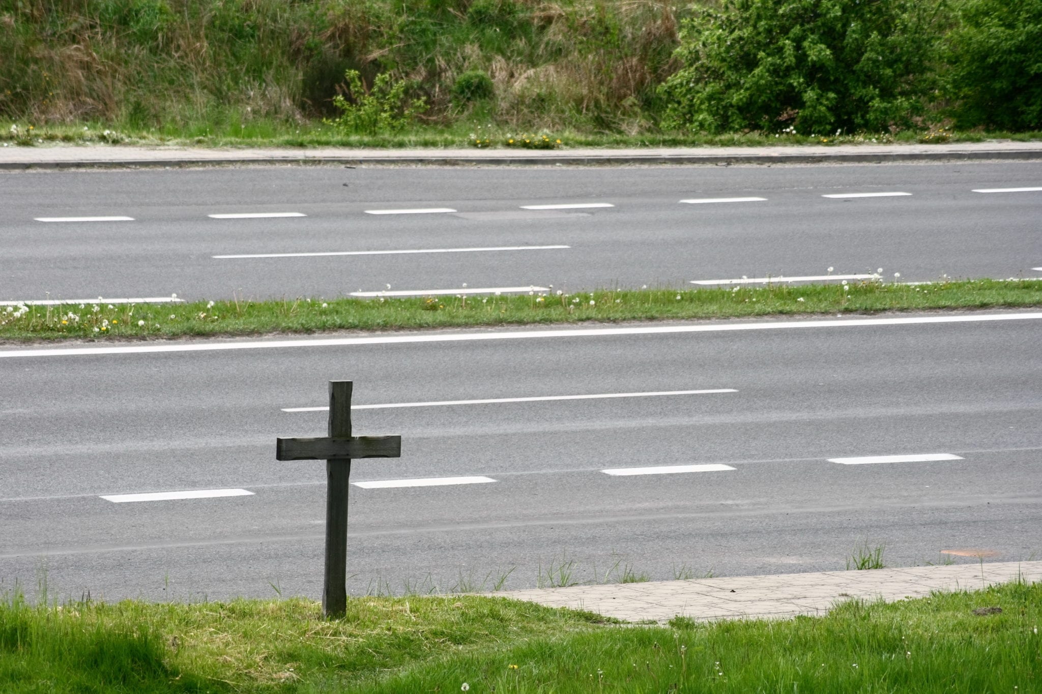 A divided multi-lane highway with green grass on each side, as well as the center median, and a wooden memoral cross standing in the grass near the road.