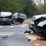 A black car and a grey car in a wreck scene on a a road with their front ends completely smashed in, appearing to have had a bad front end collision.