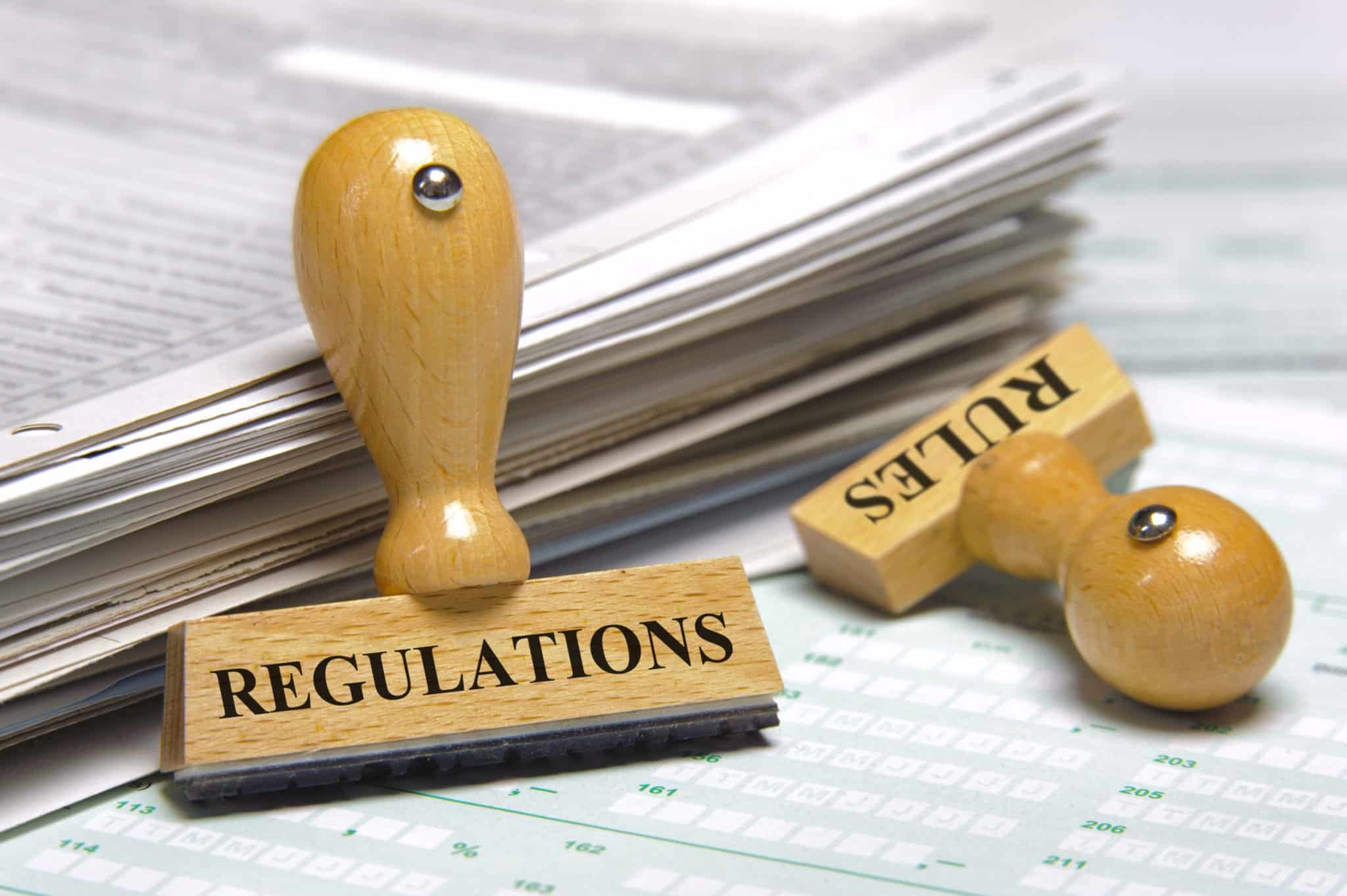 Two wooden stamps with the word REGULATIONS on one and RULES on the other, both stamps are resting on a stack of documents.