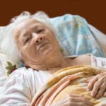 An elderly woman laying in bed with a yellow blanket over her and a stern look on her face.