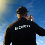 A blue sky with white clouds and a glowing yellow sun in the corner and the back of a man holding a walkie talkie, wearing a black hat and a long sleeved black uniform with the word SECURITY written in white letters across the back.