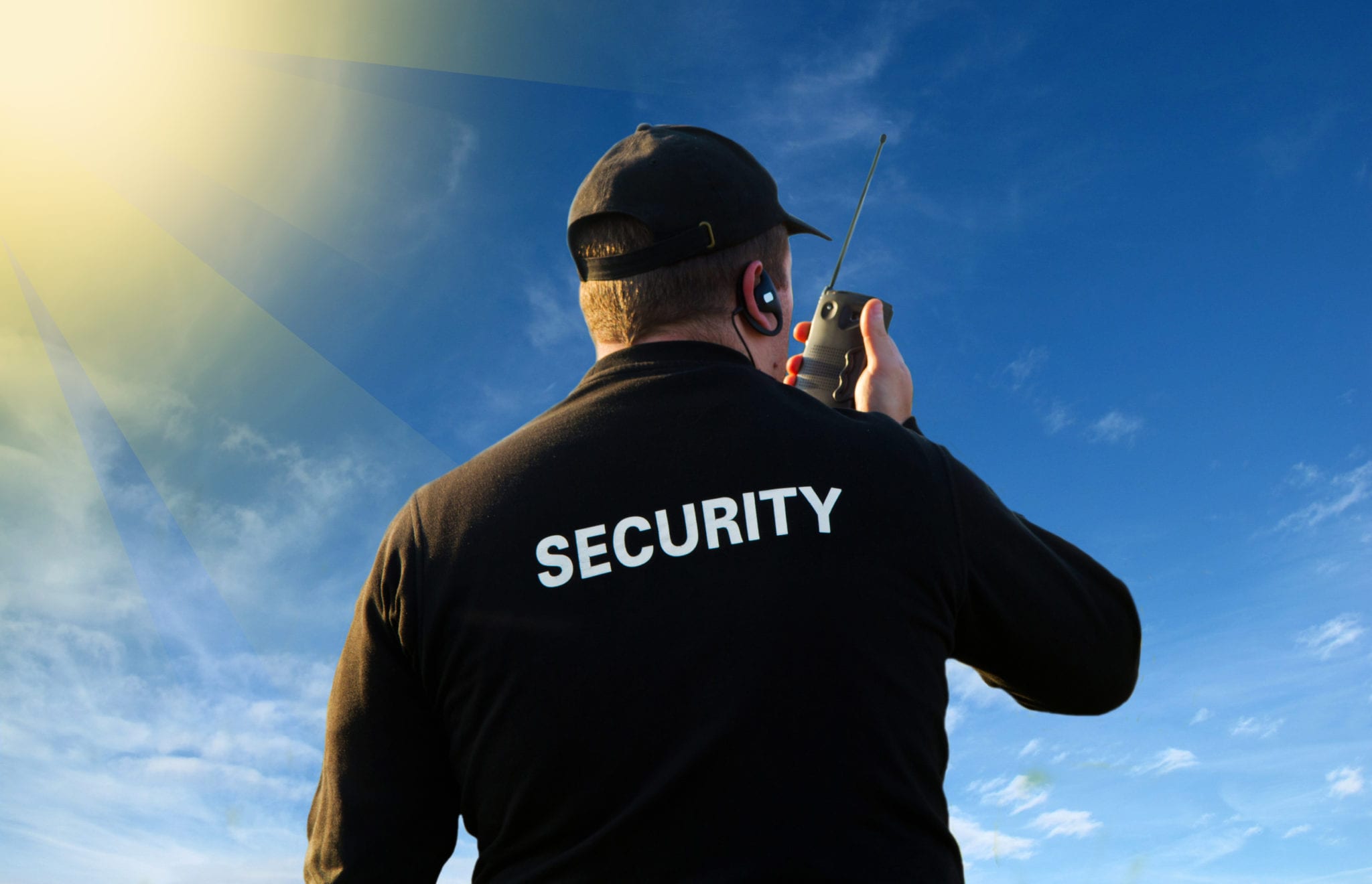 A blue sky with white clouds and a glowing yellow sun in the corner and the back of a man holding a walkie talkie, wearing a black hat and a long sleeved black uniform with the word SECURITY written in white letters across the back.