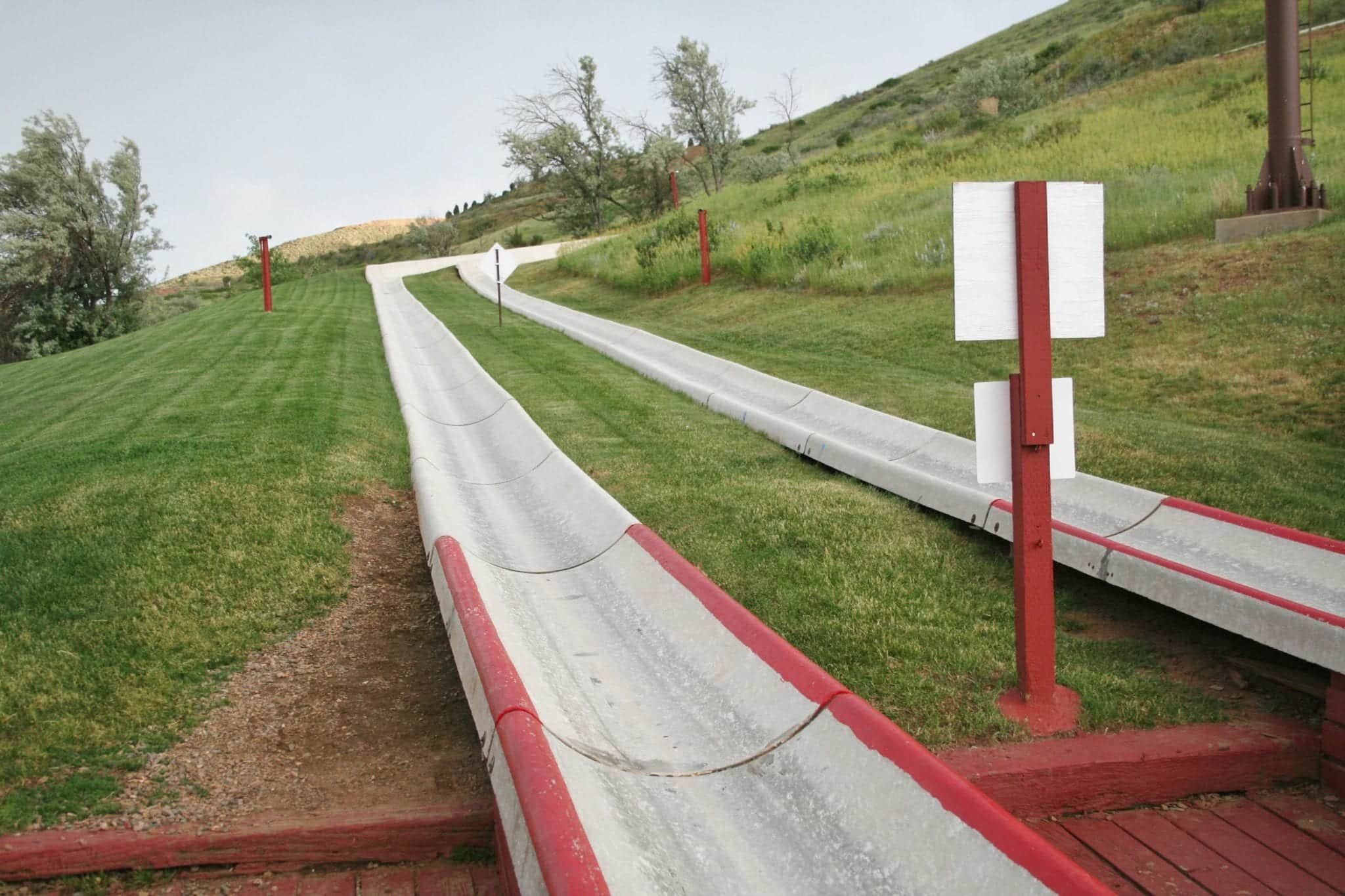 A hillside covered in short green grass with taller grass and a few trees in the background and two grey parallel slides curving down the hillside.