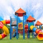 A brightly colored playground with multiple slides and platforms and a bridge, on green grass under a bright blue and white sky.