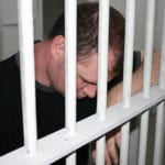 A man wearing a black t-shirt, standing in a prison cell with his hanging down and his arm propped up the white bars of the prison cell.